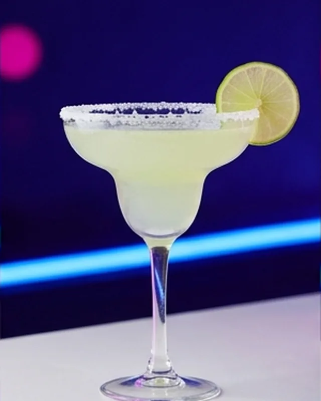 Copa de margarita con borde escarchado y rodaja de lima, sobre la barra blanca con luz azul.